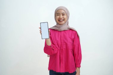 Açık pembe tesettür ve canlı mor bluz giyen neşeli genç Asyalı kadın, izole arka planda uygulamalar veya web sitesi modellemeleri için ideal beyaz ekran ile güvenli bir akıllı telefon sergiliyor