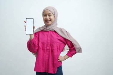 Açık pembe tesettür ve canlı mor bluz giyen neşeli genç Asyalı kadın, izole arka planda uygulamalar veya web sitesi modellemeleri için ideal beyaz ekran ile güvenli bir akıllı telefon sergiliyor