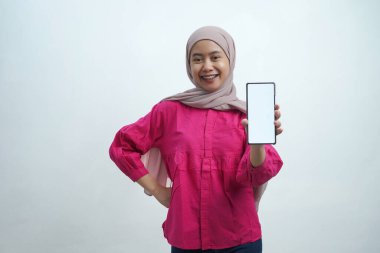 Açık pembe tesettür ve canlı mor bluz giyen neşeli genç Asyalı kadın, izole arka planda uygulamalar veya web sitesi modellemeleri için ideal beyaz ekran ile güvenli bir akıllı telefon sergiliyor