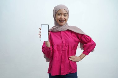 Açık pembe tesettür ve canlı mor bluz giyen neşeli genç Asyalı kadın, izole arka planda uygulamalar veya web sitesi modellemeleri için ideal beyaz ekran ile güvenli bir akıllı telefon sergiliyor
