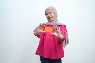 Bej tesettür ve pembe bluz giyen genç Asyalı Müslüman kadın gülümsüyor ve izole edilmiş arka planda turuncu bir kredi kartı sunuyor. Pazarlama, bankacılık ve dijital finans temaları için ideal.