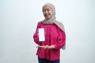 Açık pembe tesettür ve canlı mor bluz giyen neşeli genç Asyalı kadın, izole arka planda uygulamalar veya web sitesi modellemeleri için ideal beyaz ekran ile güvenli bir akıllı telefon sergiliyor