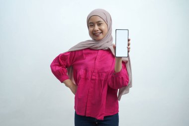 Açık pembe tesettür ve canlı mor bluz giyen neşeli genç Asyalı kadın, izole arka planda uygulamalar veya web sitesi modellemeleri için ideal beyaz ekran ile güvenli bir akıllı telefon sergiliyor