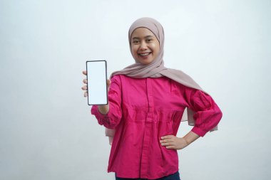 Açık pembe tesettür ve canlı mor bluz giyen neşeli genç Asyalı kadın, izole arka planda uygulamalar veya web sitesi modellemeleri için ideal beyaz ekran ile güvenli bir akıllı telefon sergiliyor