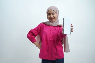 Açık pembe tesettür ve canlı mor bluz giyen neşeli genç Asyalı kadın, izole arka planda uygulamalar veya web sitesi modellemeleri için ideal beyaz ekran ile güvenli bir akıllı telefon sergiliyor