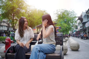 İki genç Asyalı kadın şehir bankında otururken neşeli bir sohbete girişiyor. Açık hava manzarası, hareketli bir sokak ve arka planda ağaçlar.