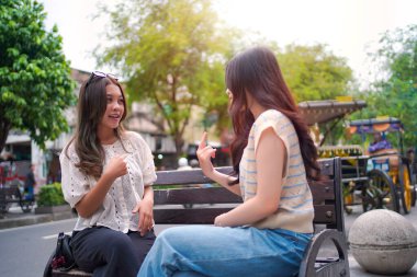 İki genç Asyalı kadın şehir bankında otururken neşeli bir sohbete girişiyor. Açık hava manzarası, hareketli bir sokak ve arka planda ağaçlar.
