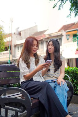İki genç Asyalı kadın şehir yaşamının ortasında akıllı bir telefona bakarken bir şehir bankında yakın oturuyorlar. Açık ve rahat bir sokak sahnesi Bağlantı ve gelişigüzel etkileşimle dolu.