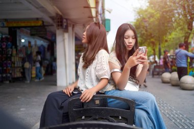 İki genç Asyalı kadın şehir yaşamının ortasında akıllı bir telefona bakarken bir şehir bankında yakın oturuyorlar. Açık ve rahat bir sokak sahnesi Bağlantı ve gelişigüzel etkileşimle dolu.