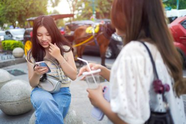 Neşeli genç Asyalı bir kadın, arkadaşı canlı bir caddenin yakınında dururken taştan bir bankta telefonunu kullanarak oturuyor. Neşe ve dijital bağlantının samimi bir kentsel anı.