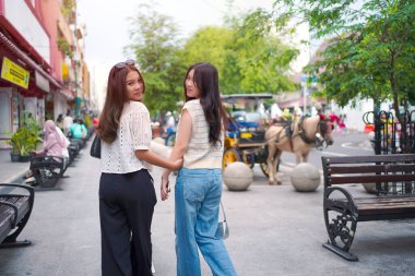 İki zarif Asyalı kadın kameraya gülümserken canlı bir şehir caddesinde çekçekçeklerle yürüyorlar..