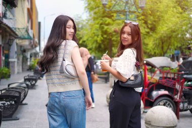 İki zarif Asyalı kadın kameraya gülümserken canlı bir şehir caddesinde çekçekçeklerle yürüyorlar..