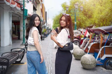 İki zarif Asyalı kadın kameraya gülümserken canlı bir şehir caddesinde çekçekçeklerle yürüyorlar..