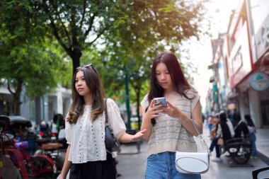 Şık genç Asyalı kadınlar canlı bir şehir caddesinde yürüyor, biri buzlu çayını yudumlarken diğeri de akıllı telefonunu kullanıyor.