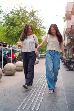 İki neşeli Asyalı kadın şehrin kaldırımında el ele yürüyorlar. Sohbet ederken ve gülümserken, şehir ortamında güneşli bir günün tadını çıkarırken buzlu bir içki içilir..