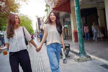 İki neşeli Asyalı kadın şehrin kaldırımında el ele yürüyorlar. Sohbet ederken ve gülümserken, şehir ortamında güneşli bir günün tadını çıkarırken buzlu bir içki içilir..