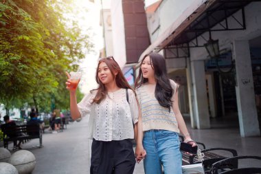 İki neşeli Asyalı kadın şehrin kaldırımında el ele yürüyorlar. Sohbet ederken ve gülümserken, şehir ortamında güneşli bir günün tadını çıkarırken buzlu bir içki içilir..