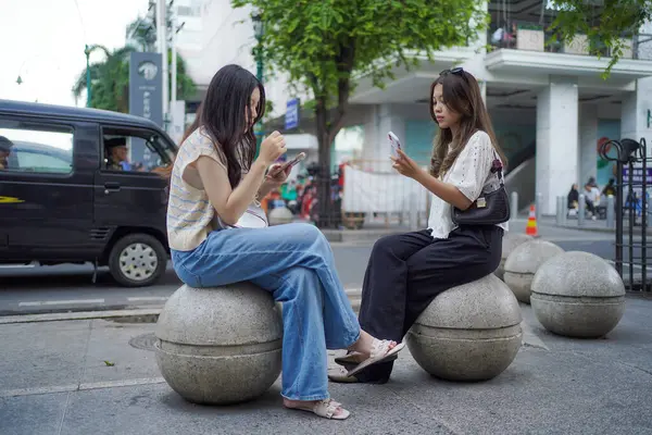 Endonezya, Yogyakarta 'da Malioboro caddesinin kenarındaki bankta otururken akıllı telefon tutan iki Asyalı kadın.