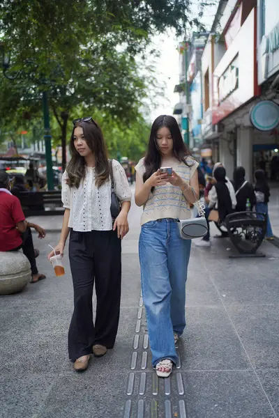 İki Asyalı kadın ellerinde cep telefonuyla sokakta yürüyor. Malioboro Caddesi, Yogyakarta, Endonezya