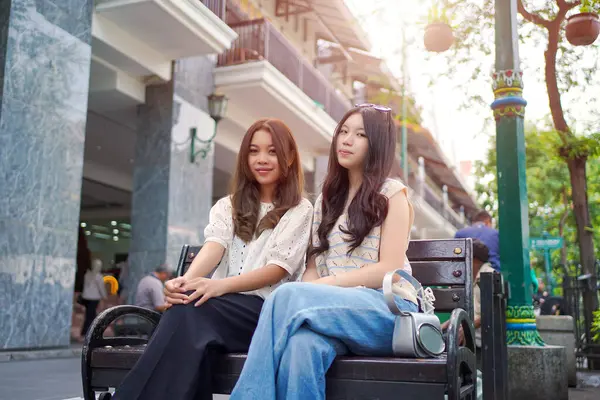 İki zarif Asyalı kadın canlı bir şehir bölgesinde, bir bankta yan yana oturuyor, gülümsüyor ve günün tadını çıkarıyorlar. Sıradan moda ve dostane atmosfer.
