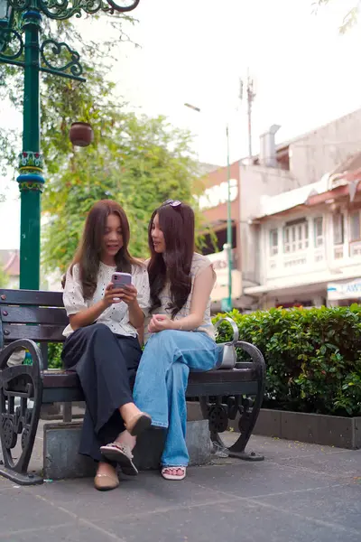 İki genç Asyalı kadın şehir yaşamının ortasında akıllı bir telefona bakarken bir şehir bankında yakın oturuyorlar. Açık ve rahat bir sokak sahnesi Bağlantı ve gelişigüzel etkileşimle dolu.