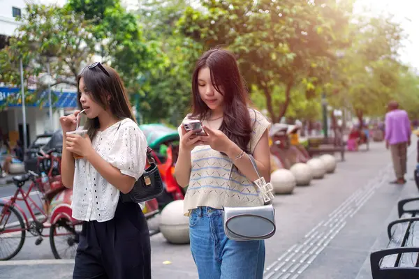 Şık genç Asyalı kadınlar canlı bir şehir caddesinde yürüyor, biri buzlu çayını yudumlarken diğeri de akıllı telefonunu kullanıyor.