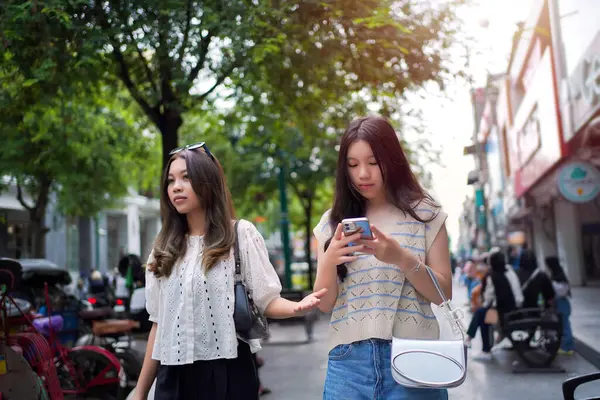 Şık genç Asyalı kadınlar canlı bir şehir caddesinde yürüyor, biri buzlu çayını yudumlarken diğeri de akıllı telefonunu kullanıyor.