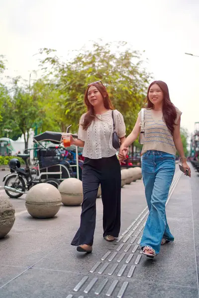 İki neşeli Asyalı kadın şehrin kaldırımında el ele yürüyorlar. Sohbet ederken ve gülümserken, şehir ortamında güneşli bir günün tadını çıkarırken buzlu bir içki içilir..