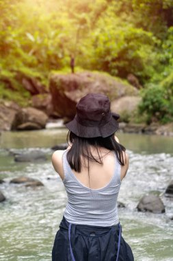 Arkaya bakan Asyalı genç bir kadın kova şapka ve atlet giyiyor, yemyeşil bir ormanda bir nehrin kenarında duruyor.