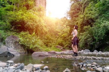 Genç Asyalı bir kadın ormanda sığ bir akarsuyun kenarında duruyor. Etrafı kaya ve doğa ile çevrili. Açık havanın tadını çıkarıyor..