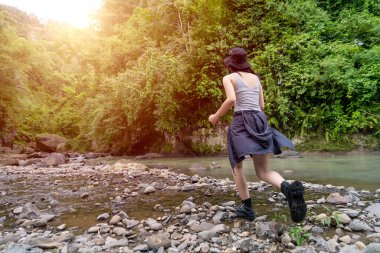 Asyalı kadın yürüyüşçünün sıradan dış görünüşü sıcak güneş ışığı altında canlı yeşil bir ormanda kayalık bir nehri geçer.