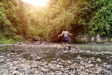 Asyalı kadın yürüyüşçünün sıradan dış görünüşü sıcak güneş ışığı altında canlı yeşil bir ormanda kayalık bir nehri geçer.
