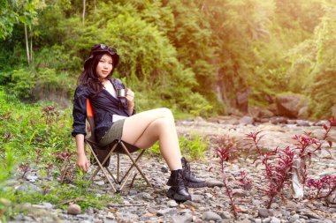 Genç Asyalı bir kadın, orman ortamında bir kamp koltuğunda oturmuş sıcak bir içkinin ve huzurlu manzaranın tadını çıkarıyor.