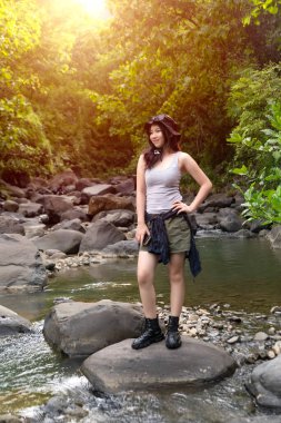 Genç Asyalı bir kadın ormanda sığ bir akarsuyun kenarında duruyor. Etrafı kaya ve doğa ile çevrili. Açık havanın tadını çıkarıyor..