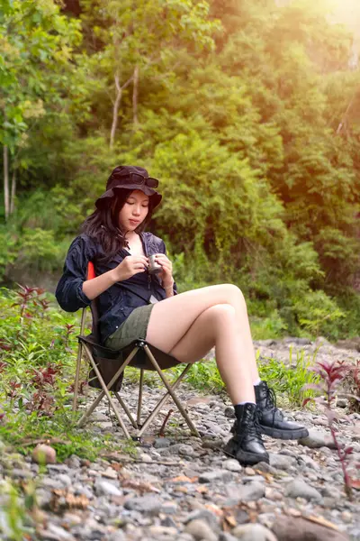 Genç Asyalı bir kadın, orman ortamında bir kamp koltuğunda oturmuş sıcak bir içkinin ve huzurlu manzaranın tadını çıkarıyor.