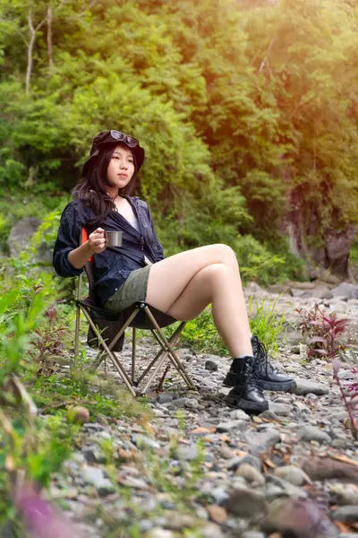 Genç Asyalı bir kadın, orman ortamında bir kamp koltuğunda oturmuş sıcak bir içkinin ve huzurlu manzaranın tadını çıkarıyor.
