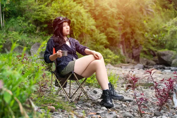 Genç Asyalı bir kadın, orman ortamında bir kamp koltuğunda oturmuş sıcak bir içkinin ve huzurlu manzaranın tadını çıkarıyor.