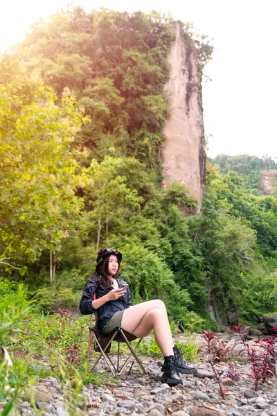 Genç Asyalı bir kadın, orman ortamında bir kamp koltuğunda oturmuş sıcak bir içkinin ve huzurlu manzaranın tadını çıkarıyor.