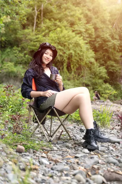 Genç Asyalı bir kadın, orman ortamında bir kamp koltuğunda oturmuş sıcak bir içkinin ve huzurlu manzaranın tadını çıkarıyor.