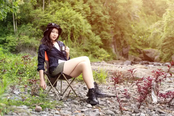 Genç Asyalı bir kadın, orman ortamında bir kamp koltuğunda oturmuş sıcak bir içkinin ve huzurlu manzaranın tadını çıkarıyor.