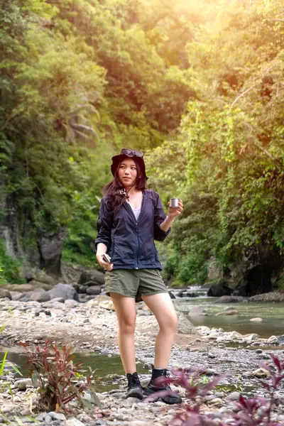 Genç Asyalı bir kadın, elinde fincanla orman yeşiliyle çevrili kayalık bir nehir kıyısında yürürken doğanın tadını çıkarıyor..