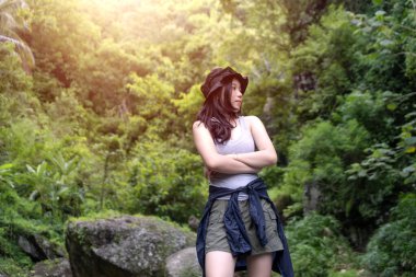 Genç Asyalı bir kadın, kolları çapraz tropikal bir ormanda, kolları bağlı, atlet bluz ve güneş şapkası takıyor, yemyeşil ve yumuşak güneş ışığıyla çevrili..