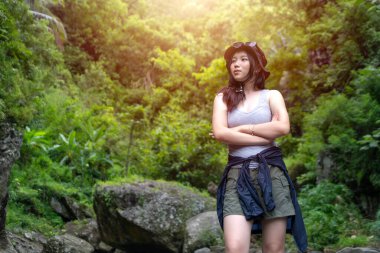 Genç Asyalı bir kadın, kolları çapraz tropikal bir ormanda, kolları bağlı, atlet bluz ve güneş şapkası takıyor, yemyeşil ve yumuşak güneş ışığıyla çevrili..