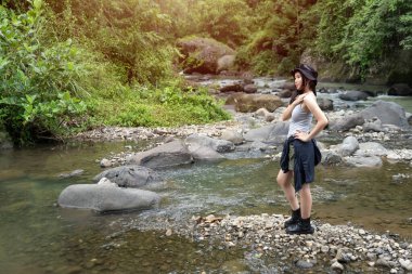 Yürüyüş kıyafetleri içinde genç bir Asyalı kadın ormanda bir nehirde kayaların üzerinde duruyor. Huzurlu bir doğa anının tadını çıkarırken gülümsüyor..