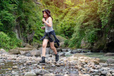 Neşeli genç Asyalı kadın canlı yeşilliklerle çevrili nehir taşlarının üzerinden koşarak doğa macerasının tadını çıkarıyor.