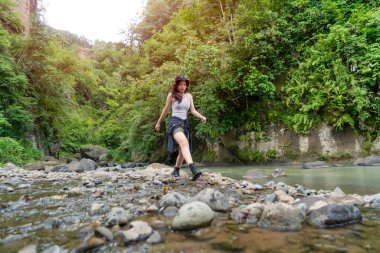 Genç Asyalı kadın yoğun tropikal bir ormanda nehir taşlarının üzerinde güven içinde yürüyüp maceralı bir yürüyüşün tadını çıkarıyor..