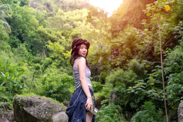 Genç Asyalı kadın gülümsüyor ve canlı yeşil bir ormanda arkasına bakıyor. Güneş şapkası ve açık hava kıyafetleri giyiyor. Yumuşak güneş ışığı altında..
