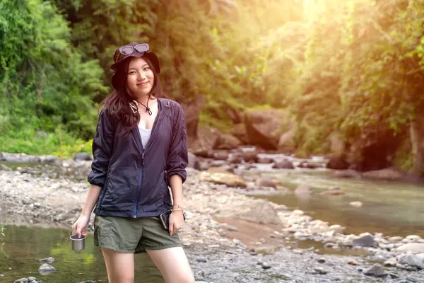 Kayalık bir nehir kıyısında elinde metal bir bardakla, gün ışığında yemyeşil ormanlarla çevrili mutlu genç Asyalı kadın..
