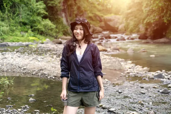 Kayalık bir nehir kıyısında elinde metal bir bardakla, gün ışığında yemyeşil ormanlarla çevrili mutlu genç Asyalı kadın..