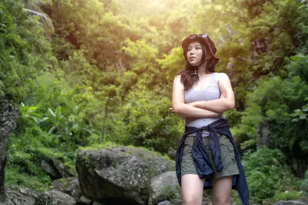 Genç Asyalı bir kadın, kolları çapraz tropikal bir ormanda, kolları bağlı, atlet bluz ve güneş şapkası takıyor, yemyeşil ve yumuşak güneş ışığıyla çevrili..